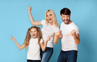 A family of three, a young girl, a woman, and a man, stand together in front of a solid blue background, all raising their fists in the air and smiling with excitement. photo