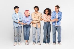 A diverse group of five young adults, all dressed casually in jeans and shirts, are standing in a row and holding hands, laughing together in a studio setting. photo