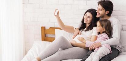 Pregnant Woman Taking Selfie With Her Husband And Daughter In Bed, Enjoying Spending Time At Home, Bonding In Bedroom photo
