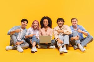 A diverse group of five young adults, all smiling and looking at the camera, are sitting on a yellow background. They are holding a laptop and pointing at it, as if showing it off to someone photo