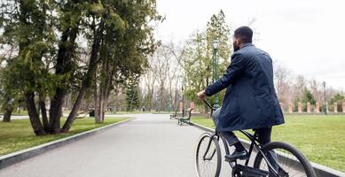 Black businessman is cycling near a green public park. The mood is dynamic and calm, showing a seamless transition between urban work life and natural relaxation. photo