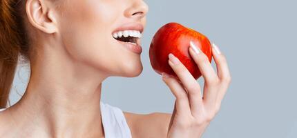 A woman is taking a bite out of a red apple, showing off her white teeth. The apple is held in her right hand, and the background is a soft grey. photo