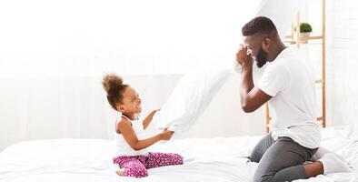A little girl with curly hair sits on a white bed, laughing and holding a white pillow while her father, who is kneeling in front of her, swings a white pillow towards her in a playful pillow fight. photo