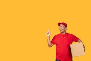 A cheerful guy delivery person dressed in a red uniform points upward with a confident smile while holding a package. The background is a vibrant yellow, copy space photo