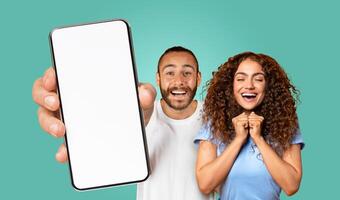 A young couple is standing in front of a turquoise background. The man is holding a smartphone with a blank screen in front of him, with his finger pointing at the screen. photo