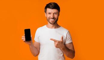 A man in a white t-shirt holds a black smartphone with a blank screen and points to it with a smile, standing against an orange background. photo