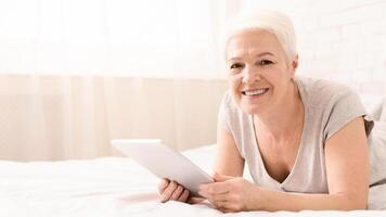 Elderly woman is reclining on a bed while holding a tablet in her hands. She appears engrossed in the devices screen, possibly reading or watching something, copy space photo