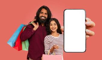 A smiling Indian couple stands in front of a pink background, holding shopping bags and a large smartphone with a blank screen. photo