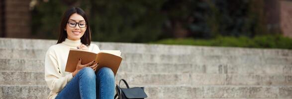 A young Asian woman wearing glasses sits on stone steps in front of a campus building. She is writing in a notebook while looking down at the page, copy space photo