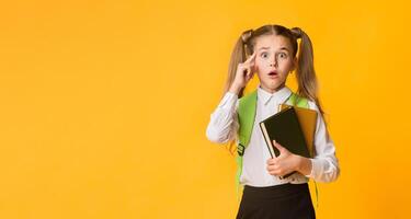 Oops. Elementary Student Girl Looking At Camera Holding Books On Yellow Background, Empty Space photo