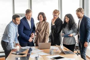 A group of diverse business professionals in formal attire gather around a table in a modern office space. They are engaged in a collaborative meeting, discussing documents and a laptop photo