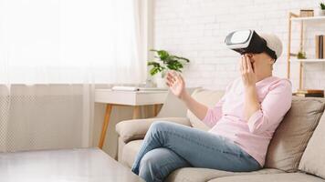 Senior woman is seated on a couch, fully immersed in a virtual reality experience as she wears a headset. Her hands are likely interacting with unseen objects in the virtual world photo
