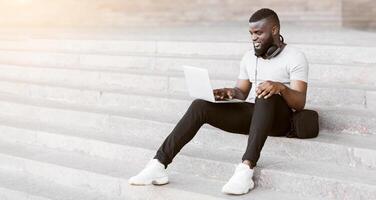 A man sits on a set of concrete stairs in an urban environment, working on a laptop while smiling. photo