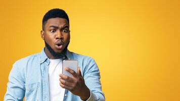 Unexpected message. Astonished african american man looking at smartphone screen in shock, standing over yellow background with copy space photo