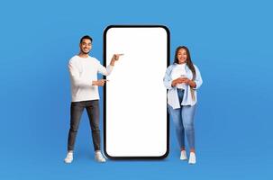Multiethnic man and woman are standing side by side next to large phone, looking at the screen and appearing to have conversation. The phone is significantly bigger than them, emphasizing its size. photo
