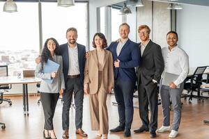 A group of six diverse business professionals stand together in a modern office setting. They are all smiling and appear to be in a good mood. The team is comprised of two women and four men photo