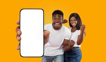 A happy black couple stand in front of a bright yellow background. The man is holding a smartphone with a blank white screen, pointing his finger at it with a wide smile photo