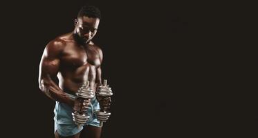 A muscular man with a bare chest and blue shorts lifts weights in front of a black background. He is looking down at his hands. photo