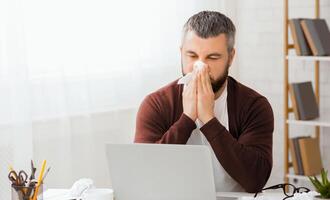 Sick man sitting in front of a laptop is blowing his nose into a tissue. He appears to be dealing with a cold or allergies while using the computer. photo