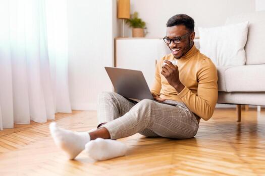Freelance Lifestyle. Relaxed Afro Guy Working On Laptop Drinking Coffee Sitting On Floor Indoor. Selective Focus photo