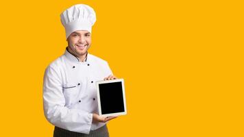 Professional Cook Guy Showing Empty Tablet Screen Standing In Studio Over Yellow Background. Panorama, Mockup photo