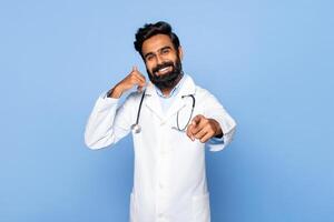 Indian doctor with a beard is smiling and making a call me gesture with his left hand while pointing his right index finger forward. He is standing in front of a blue background. photo