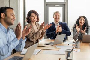 A group of diverse colleagues is sitting around a table in a modern office space. They are clapping and smiling, clearly pleased with the outcome of a successful meeting photo