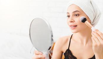 A woman is using a mirror to apply makeup with a brush in a sunny room. She is wearing a bathrobe and a towel on her head, suggesting its part of her morning routine. photo