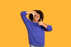 A young man with curly dark hair is wearing a blue sweatshirt and is standing against a yellow background. He is looking directly at the camera and smiling while making a framing gesture photo