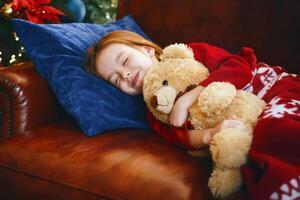 Cute little girl sleeping next to illuminated Christmas tree, dreaming about christmas miracles, hugging teddy bear, free space photo