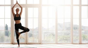 Young sporty woman doing yoga, practicing tree pose over big panoramic window, morning exercising, copy space photo