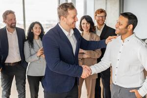 A businessman in a blue suit shakes the hand of a new employee while colleagues stand around them. All of the individuals are smiling and seem to be enjoying the moment. photo