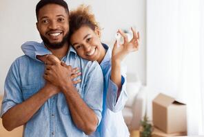 House Ownership. Smiling Afro Spouses Showing Key To Camera Embracing Standing In New Home After Relocation. Selective Focus photo