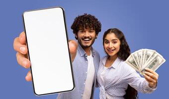 A young Indian couple is holding a smartphone with a blank screen and a fan of money, both facing the camera and smiling. photo