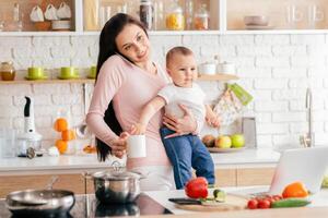 A mother is holding her baby while talking on the phone, juggling a coffee mug, and surrounded by fresh vegetables in a modern kitchen during the morning. photo