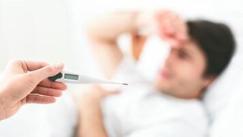 A hand holds a digital thermometer while pointing it at a man lying in bed. The man is out of focus and appears to be sick. photo