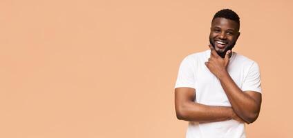 A young African American man in a white t-shirt smiles brightly while standing against a peach colored background. He has a thoughtful expression and his right hand is resting on his chin, copy space photo