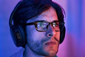 A close-up portrait of a young man wearing glasses and headphones. He is illuminated by blue and pink lighting, and his expression is serious and focused photo