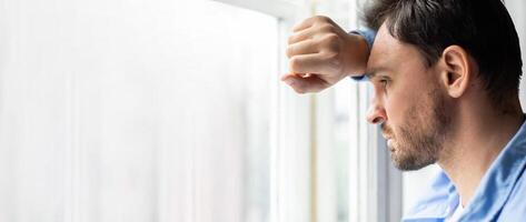 A man leans his forearm against the window, lost in thought as raindrops slide down the glass. The indoor setting captures a contemplative moment on a cloudy day. photo