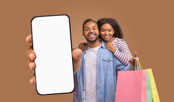A smiling black couple with shopping bags stands in front of a brown background. The man is holding a large smartphone with a blank screen, pointing to the device with his index finger photo