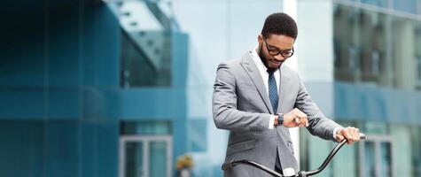 An executive black guy on a bicycle is checking his watch in a cityscape. The mood is lively and current, highlighting the importance of punctuality and an active, professional lifestyle. photo