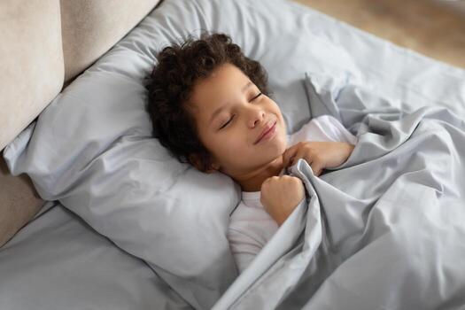 un joven africano americano chico pacíficamente duerme en un cama, cubierto por un frazada. su ojos son cerrado como él descansa cómodamente en un acogedor ambiente. foto