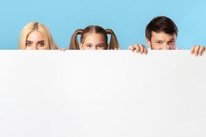 A family, consisting of a mother, father, and daughter, are peeking over a large white sign. They are smiling and looking at the camera. The background is a light blue color. photo