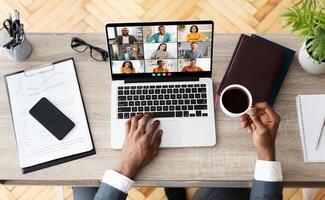 Black businessman sits at his desk, working from home. He is engaged in a conference with colleagues, visible on his laptop screen, view above photo