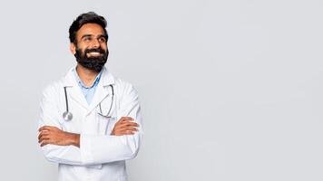 A smiling Indian man doctor wearing a white lab coat and stethoscope stands in front of a plain white background, arms crossed, looking at copy space photo