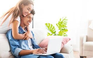 A teen guy and girl are relaxing on a couch. The girl is hugging the guy from behind, and he is looking at the laptop on his lap. The couple seems happy and comfortable in each others company. photo