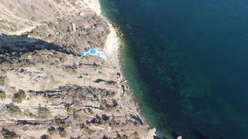 Aerial drone view of a man flying a white and blue paraglider over a hill and trees to the sea waves near the rocks. Active paraglider flight over the seascape with clear skies at suset. Extreme sport video