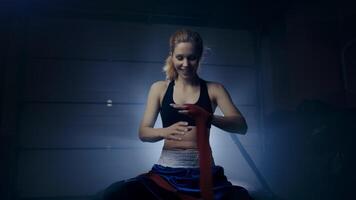 Woman boxer preparing for a fight wraps bandages around her hands in a dark room with a light behind her. video