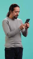 Vertical Man holding mobile phone, browsing betting website, celebrating after winning money, isolated over studio background. Cheerful person feeling happy while scrolling on smartphone, camera A video
