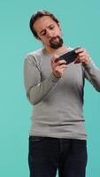 Vertical Delighted man playing videogames on smartphone, celebrating after being victorious against opponents online, studio backdrop. Player thrilled about winning game on mobile phone, camera A video